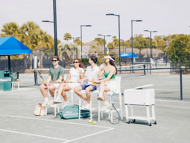 Four people sit on tall chairs at a sunny outdoor tennis court, with bags and a cooler nearby, watching something off-camera.
