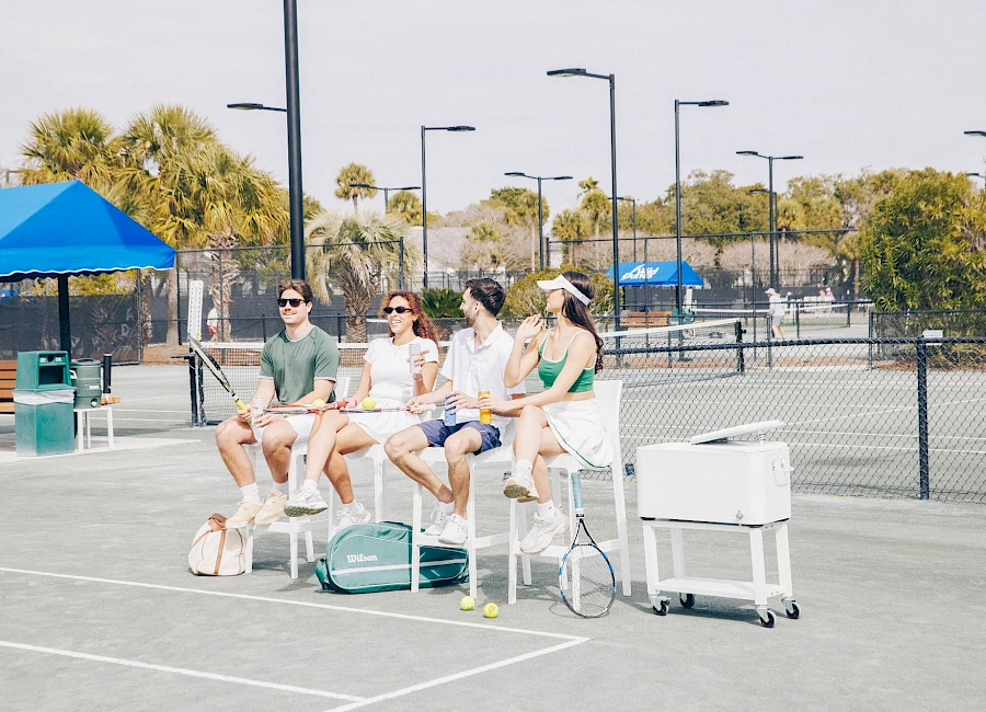 Four people sit on tall chairs at a sunny outdoor tennis court, with bags and a cooler nearby, watching something off-camera.
