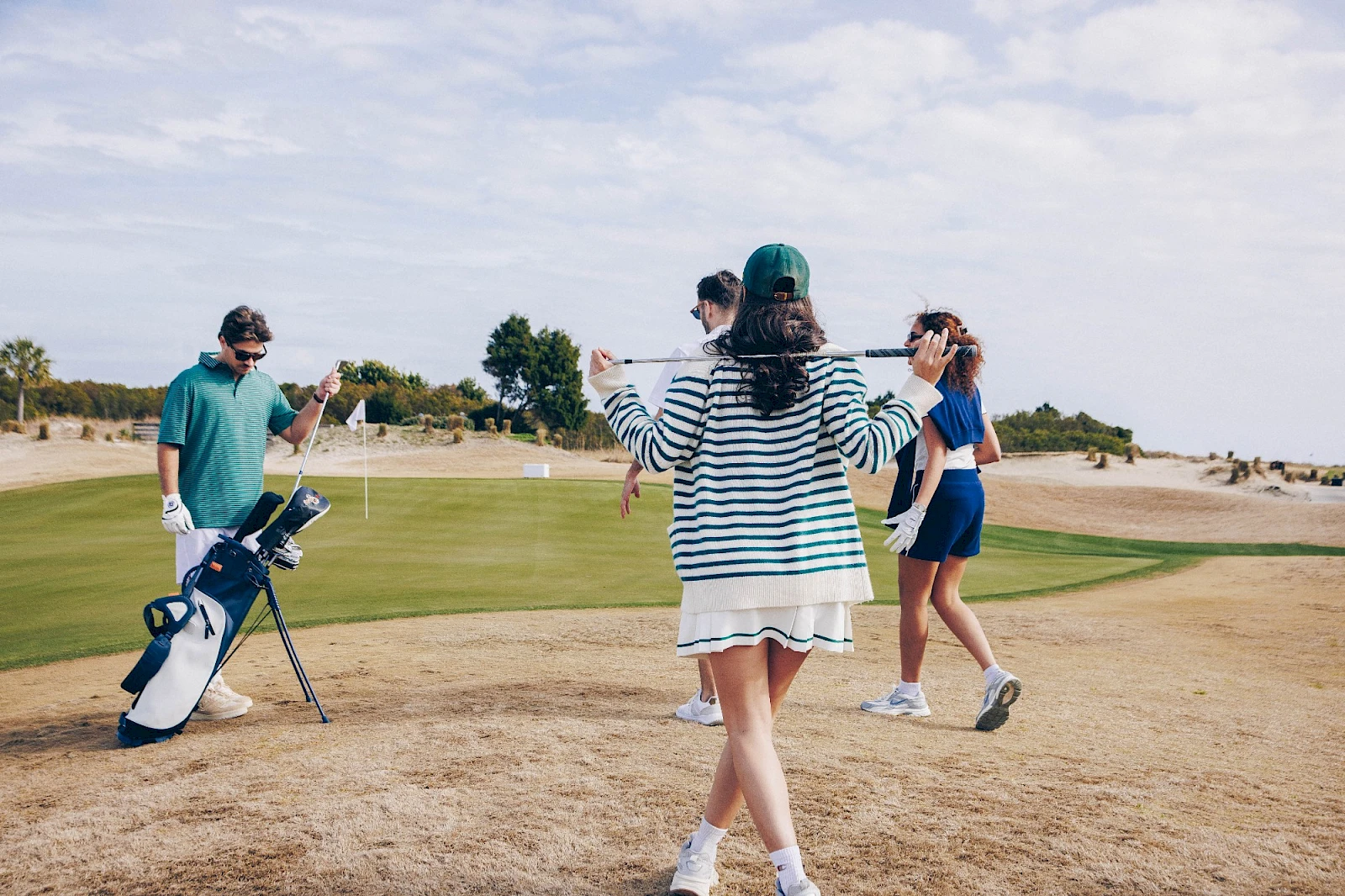 People playing golf on a sunny course; a person in striped dress faces away with arms out, another in blue shorts walks, a golfer with bag nearby, sandy terrain.