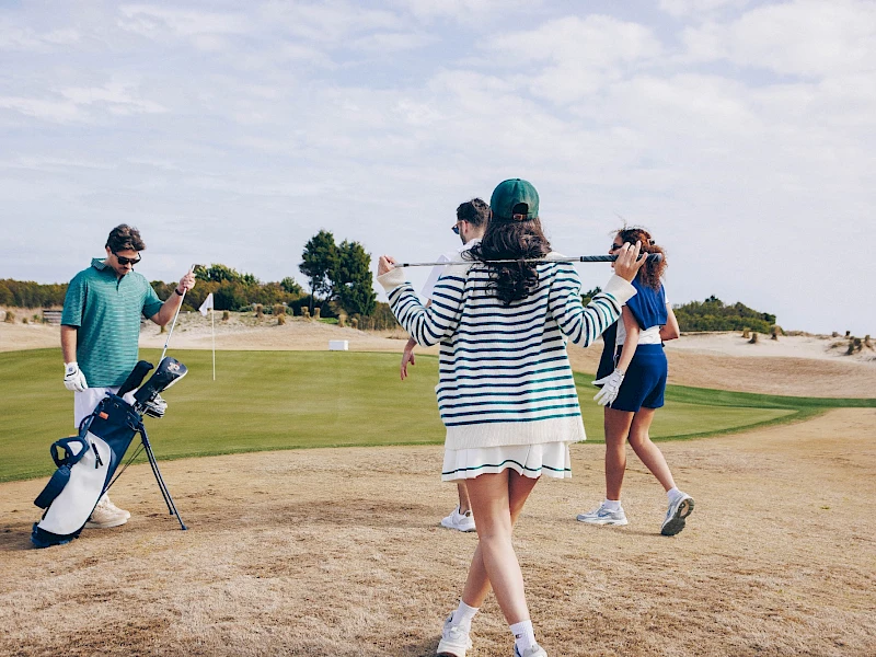 People playing golf on a sunny course; a person in striped dress faces away with arms out, another in blue shorts walks, a golfer with bag nearby, sandy terrain.