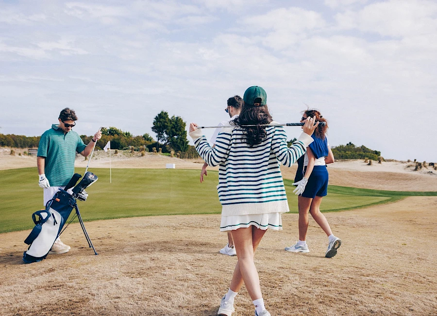 People playing golf on a sunny course; a person in striped dress faces away with arms out, another in blue shorts walks, a golfer with bag nearby, sandy terrain.