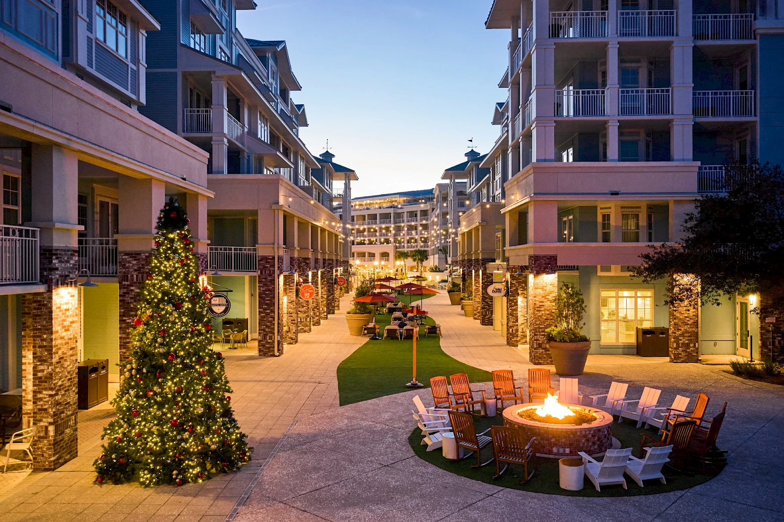 Outdoor shopping plaza at dusk with holiday lights, a lit fire pit circle, chairs, Christmas tree, and modern apartment buildings lining a pedestrian street.