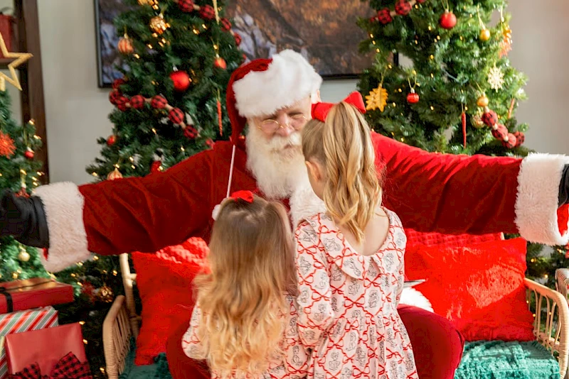 A cozy scene with a family meeting Santa: two young girls hug Santa, festive lights and Christmas trees surround them, gifts nearby, warm holiday vibes.