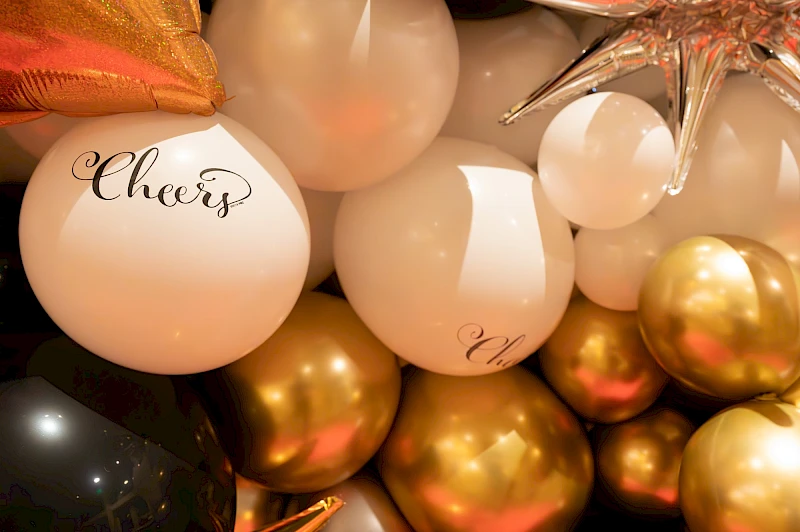 A festive cluster of white, gold, and rose balloons with cursive “Cheers” text, creating a celebratory party vibe.