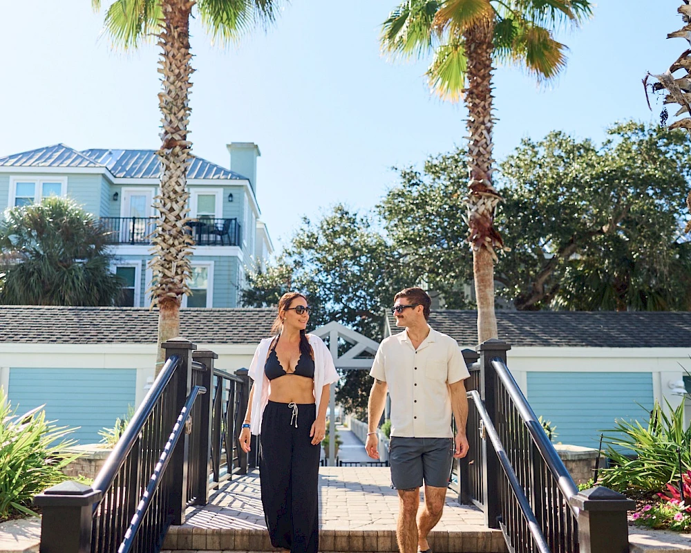 Two people walk up a set of outdoor stairs between palm trees on a sunny day, smiling and chatting as they head toward a cozy building.