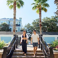 Two people walk up a set of outdoor stairs between palm trees on a sunny day, smiling and chatting as they head toward a cozy building.