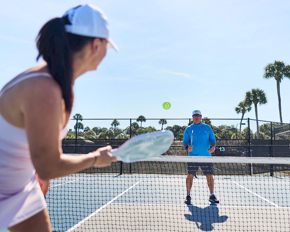 Two people play tennis on a sunny outdoor court, with a woman in the foreground about to hit the ball and a man standing at the net.