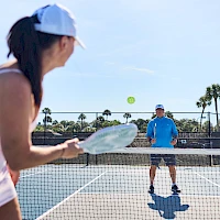 Two people play tennis on a sunny outdoor court, with a woman in the foreground about to hit the ball and a man standing at the net.