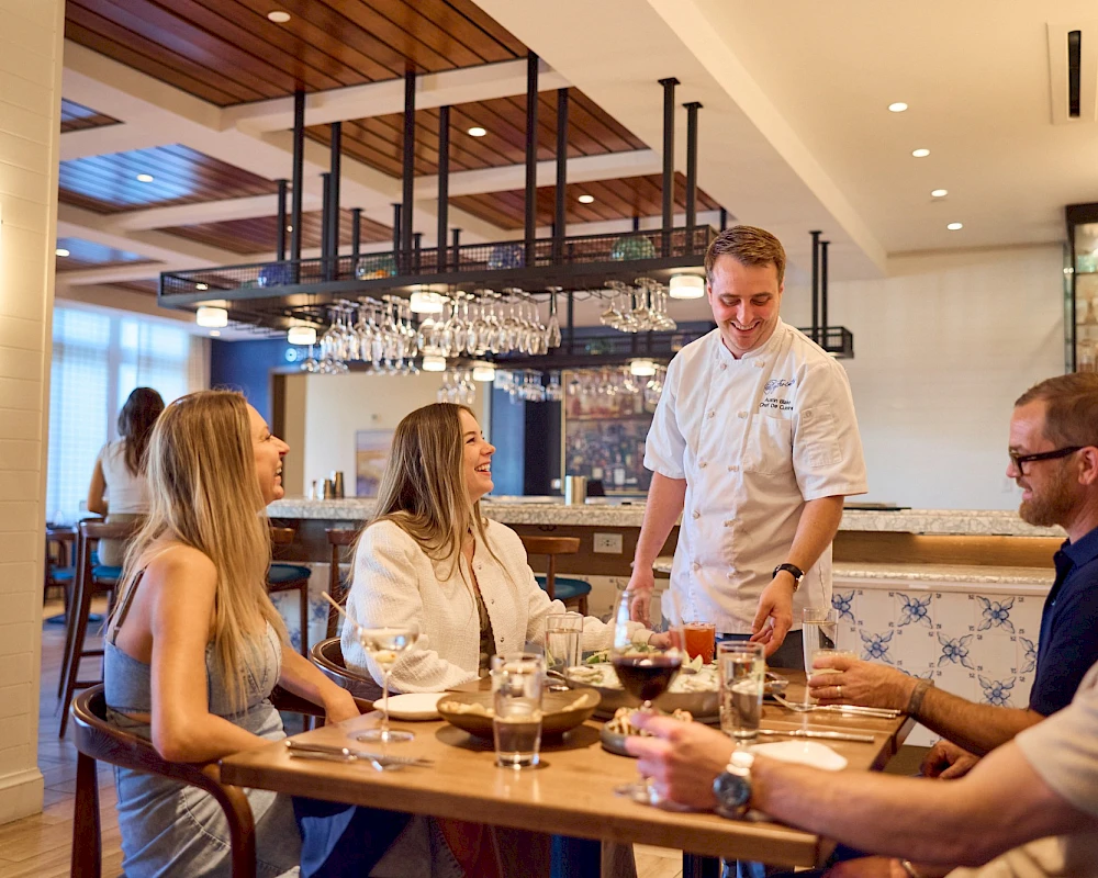 A waiter serves a group at a wooden table in a modern, well-lit restaurant, with wine glasses and engaged friends.