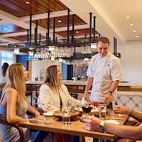 A waiter serves a group at a wooden table in a modern, well-lit restaurant, with wine glasses and engaged friends.