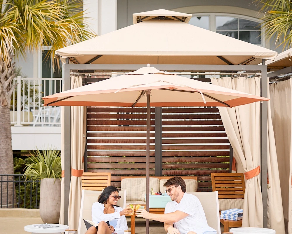 Two people chat and laugh under large beach umbrellas on lounge chairs by a poolside cabana, enjoying a sunny, relaxed moment.