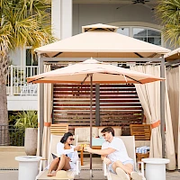 Two people chat and laugh under large beach umbrellas on lounge chairs by a poolside cabana, enjoying a sunny, relaxed moment.
