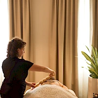 A massage therapist works on a client lying face down on a table, in a dimly lit room with beige curtains and a plant nearby, creating a calm spa atmosphere.