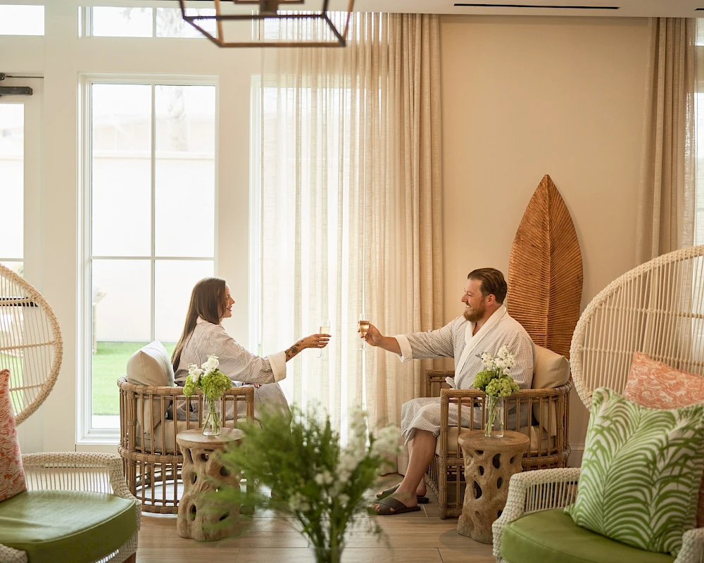 Two people sit on wicker chairs in a bright, airy living room, toasting with drinks as plants and cushions decorate the space.