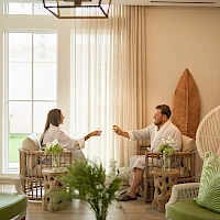 Two people sit on wicker chairs in a bright, airy living room, toasting with drinks as plants and cushions decorate the space.