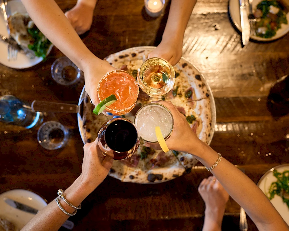 A top-down view of friends clinking colorful cocktails over a shared plate, celebrating with drinks and food on a rustic wooden table.