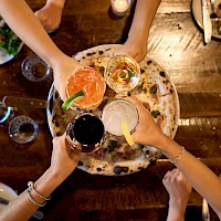A top-down view of friends clinking colorful cocktails over a shared plate, celebrating with drinks and food on a rustic wooden table.