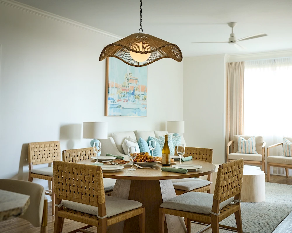 A stylish dining area in a bright, airy living space with a round wooden table set for a meal, woven chairs, and a wicker pendant light hanging above. It reads as a cozy, coastal-inspired room with a soft neutral palette and light-colored furnishings.