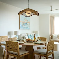 A stylish dining area in a bright, airy living space with a round wooden table set for a meal, woven chairs, and a wicker pendant light hanging above. It reads as a cozy, coastal-inspired room with a soft neutral palette and light-colored furnishings.