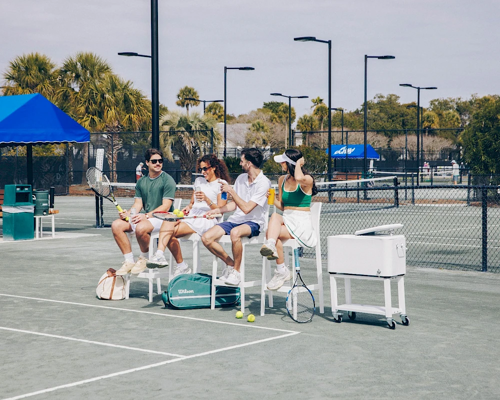 A group of people sits on a bench at a tennis court, chatting and relaxing under tents, with coolers and bags nearby.