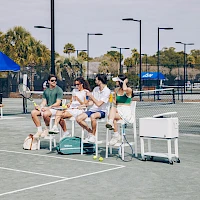 A group of people sits on a bench at a tennis court, chatting and relaxing under tents, with coolers and bags nearby.