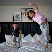Two kids jump and play on a bed in a hotel room, one in mid-air and the other landing, both smiling and energetic.