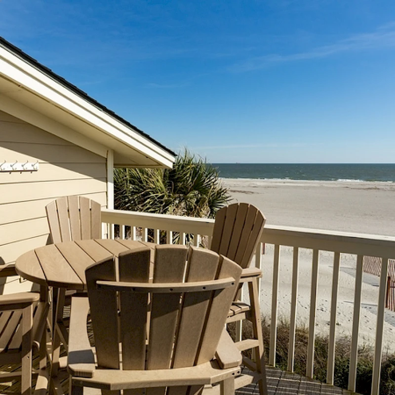 A sunny beachside balcony with tan wooden chairs around a round table, overlooking a white-sand shore and calm blue ocean.