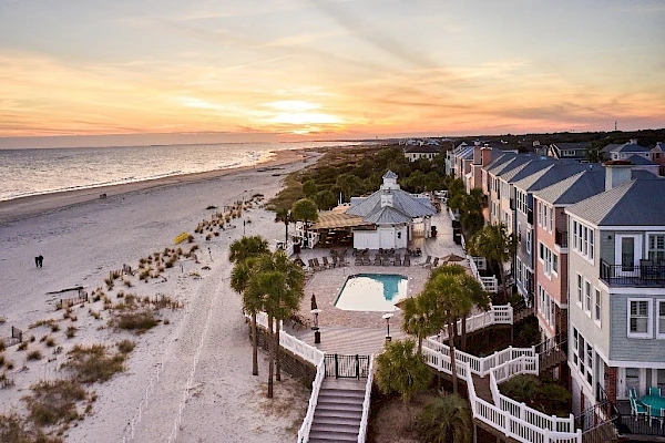 Aerial view of a coastal beach town at sunset, with a sandy shoreline, palm trees, and a pool area beside pastel beachfront houses.