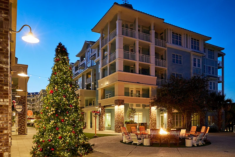 A festive outdoor plaza with a lit Christmas tree and seating around a glowing fire pit in front of a modern apartment complex. (140 chars)