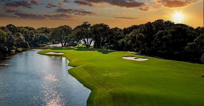 A scenic golf course along a waterway at sunset, with green fairways, sand bunkers, trees lining the edges, and shimmering water nearby.