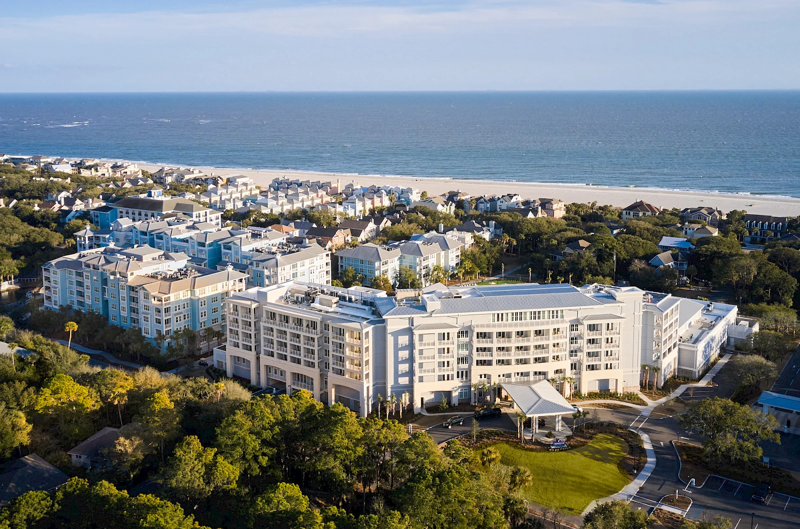Aerial view of a coastal town with white apartment buildings, palm trees, and a sandy beach meeting the blue ocean, sunny and calm day.