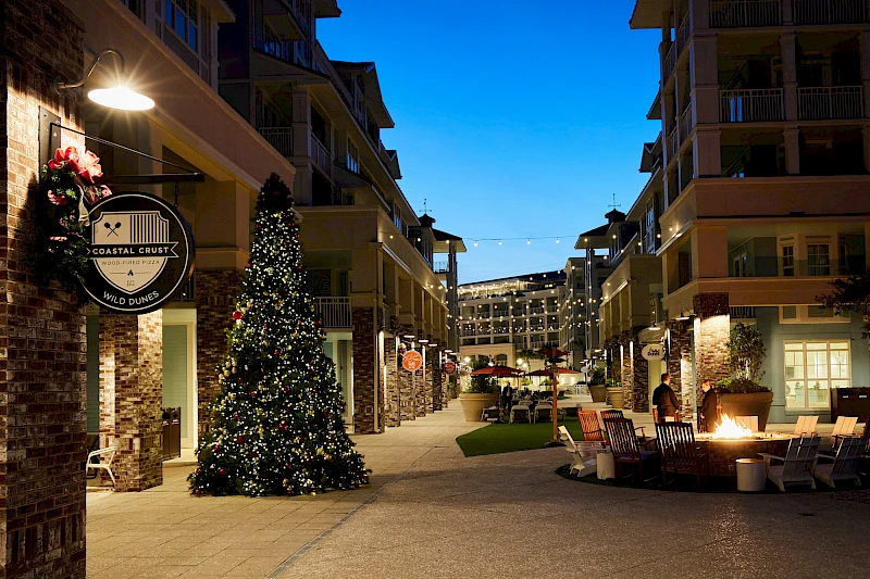 A festive outdoor shopping street at dusk with a large decorated Christmas tree, string lights, benches, and storefronts warmly lit, inviting strolls.