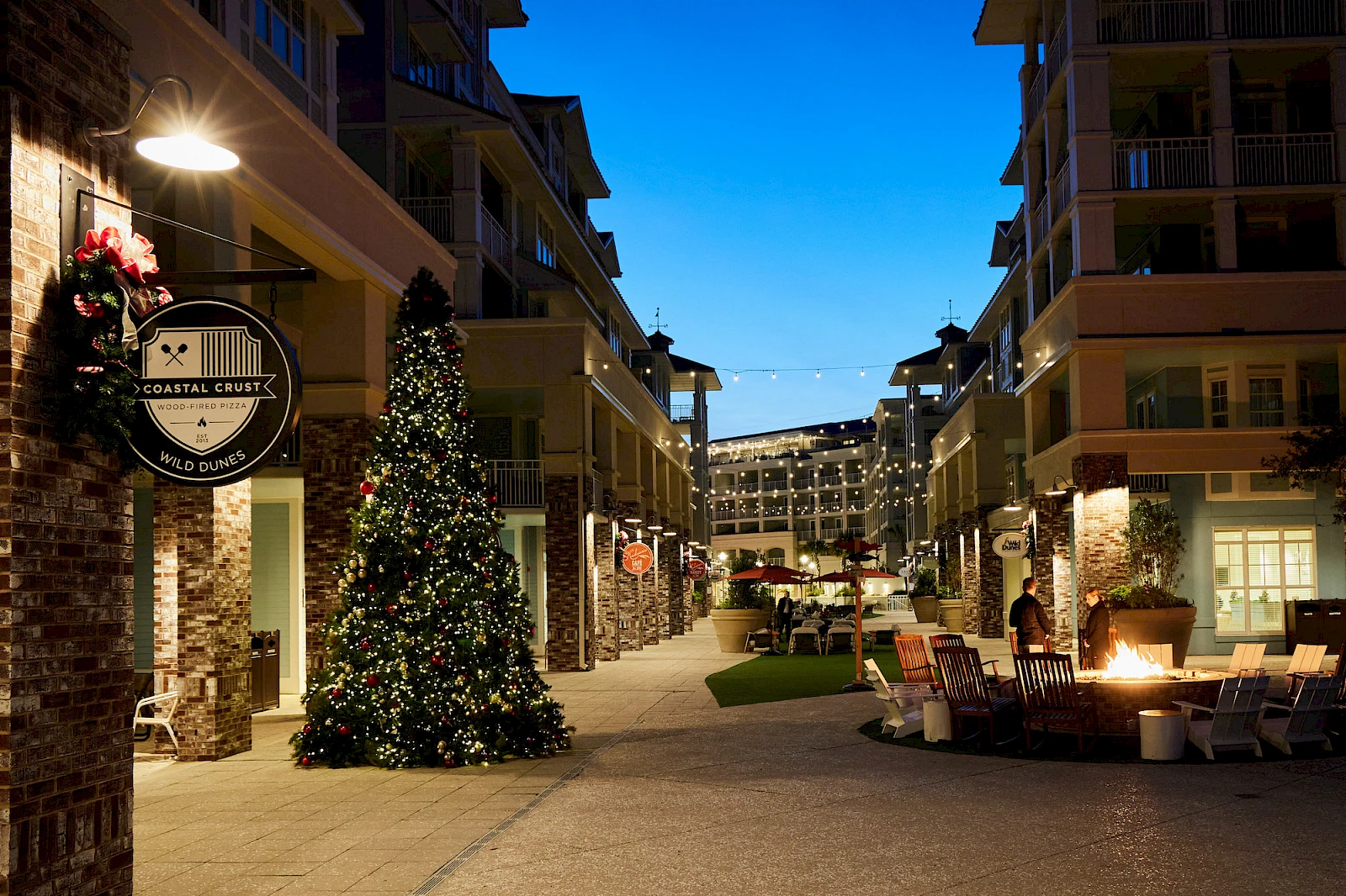 A festive outdoor shopping district at dusk with string lights, a lit Christmas tree, storefronts, and seating areas along a pedestrian street.