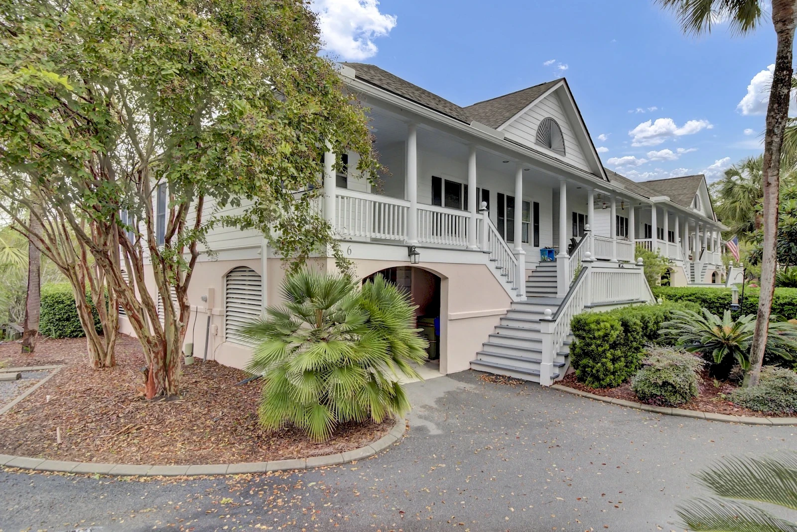 A row of light gray townhomes with a covered front porch, stairs to the second level, lush landscaping, palm trees, and a curving driveway.