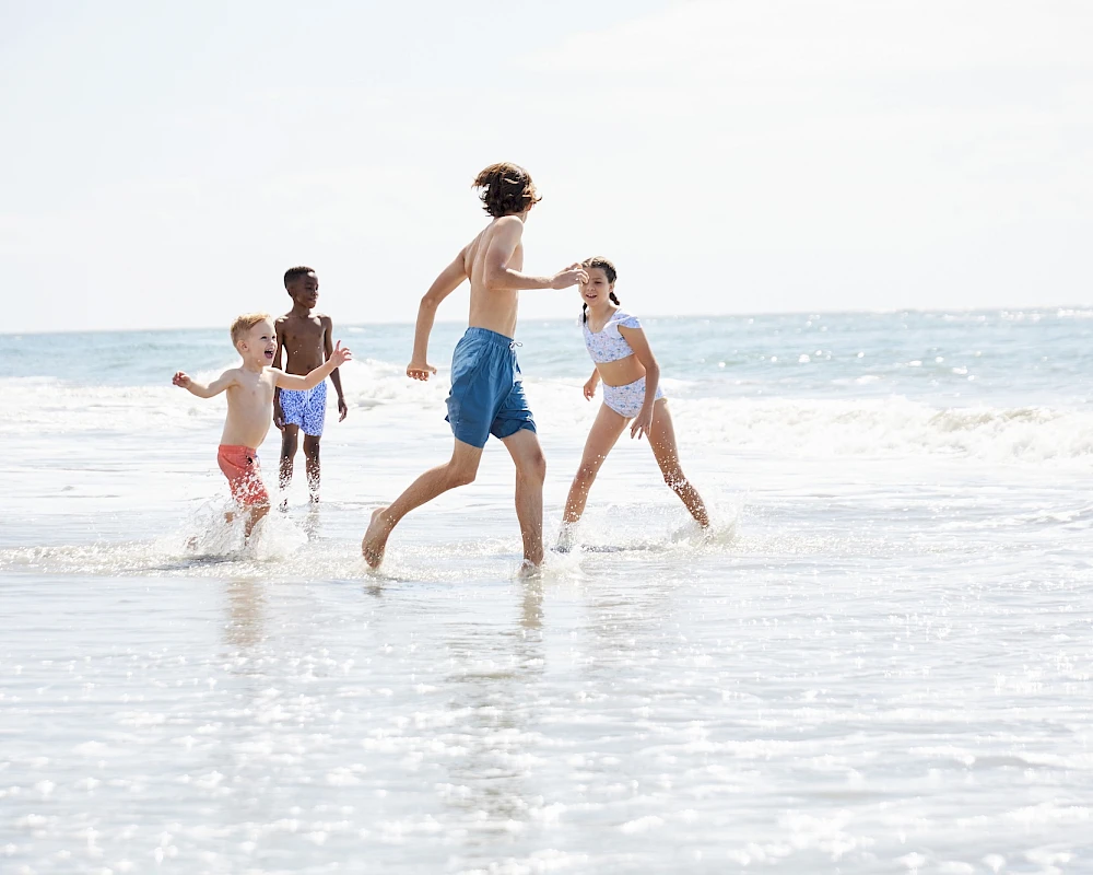 A family plays at the beach: adults and children run near the shore, waves splash, and the sunlit sea stretches to the horizon.