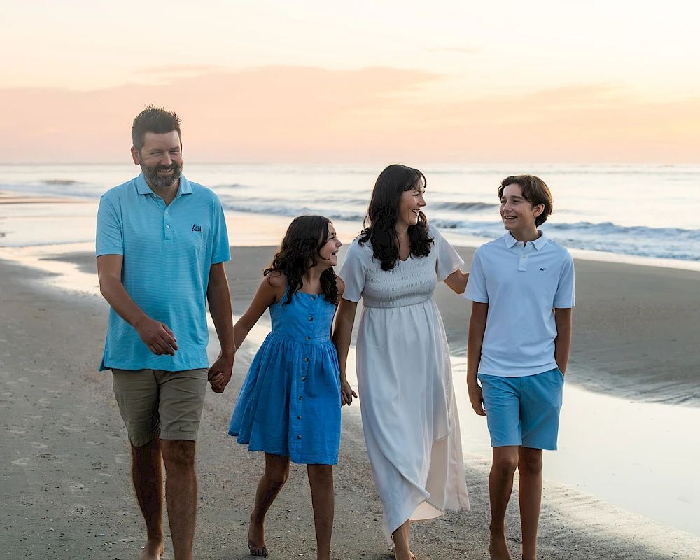 A family of four walks along a beach at sunset, holding hands and smiling, with a calm sea and warm sky in the background.