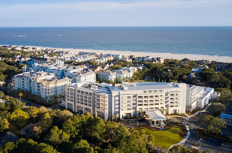 A coastal resort town with a large white apartment complex, lush greenery, and a sandy beach along the ocean.
