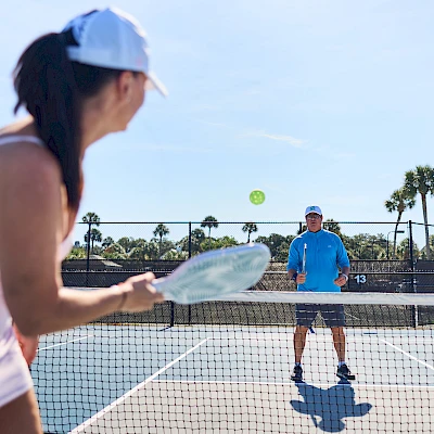Two people play tennis on an outdoor court; a woman in a white outfit serves near the net while a man in blue waits for the return under sunny skies.