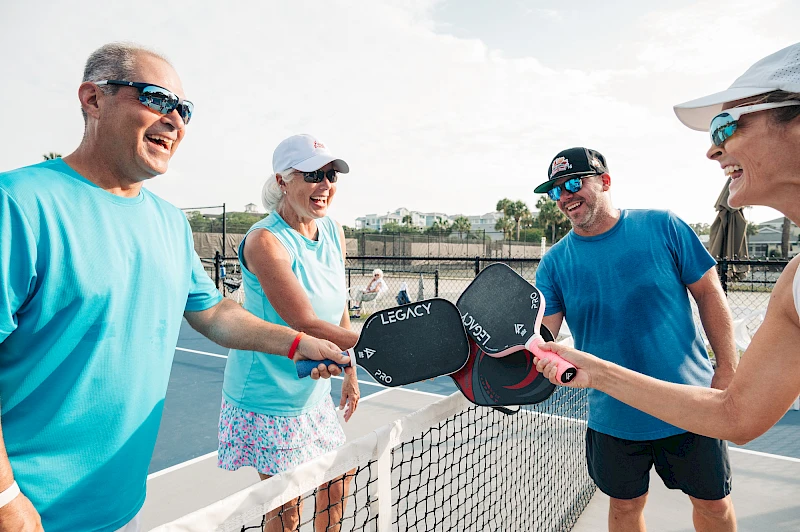 Five friends on a pickleball court high-five after a game, smiling and holding paddles, sunny day, casual athletic outfits.