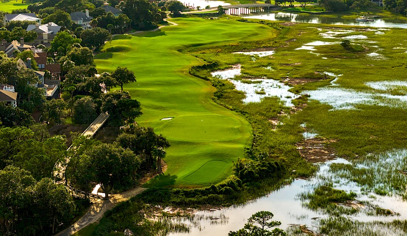 A lush golf course bordered by water and trees, with green fairways winding between residential areas and marshy wetlands along the edge.