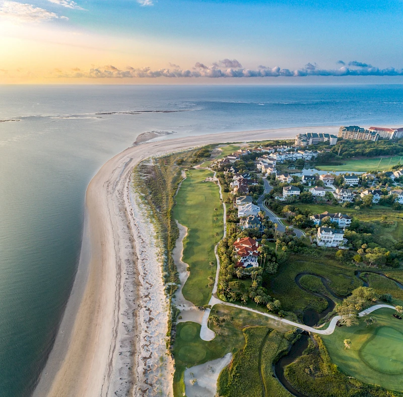 An aerial view of a coastal resort town: sandy beach, calm sea, lush villas, winding roads, and green golf courses along the shoreline.