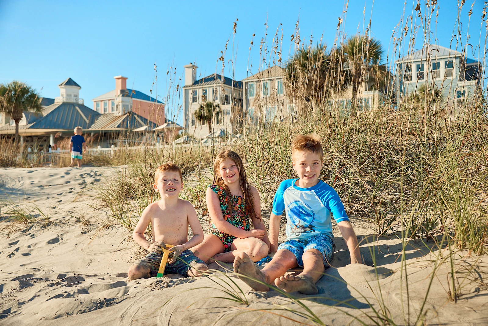 Three kids sit on the sandy beach near tall grass and dunes, with houses in the background on a sunny day, smiling and enjoying the shore.