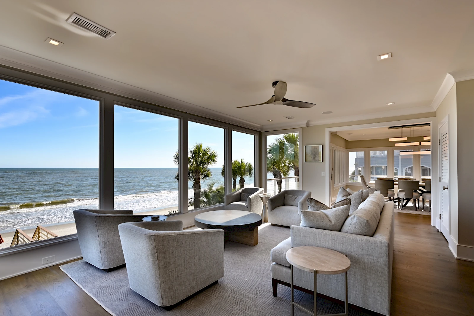 A bright beachfront living room with large windows, grey sofas, round coffee tables, palm trees outside, and a view of the ocean, ceiling fan.
