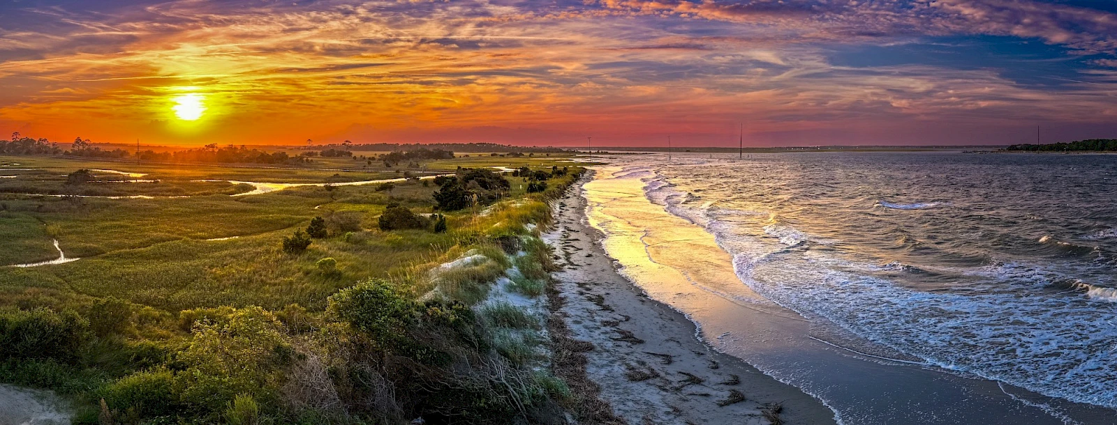 Sunset over a tranquil coastline with golden light reflecting on gentle waves, marshy greens, and a distant horizon of calm water.