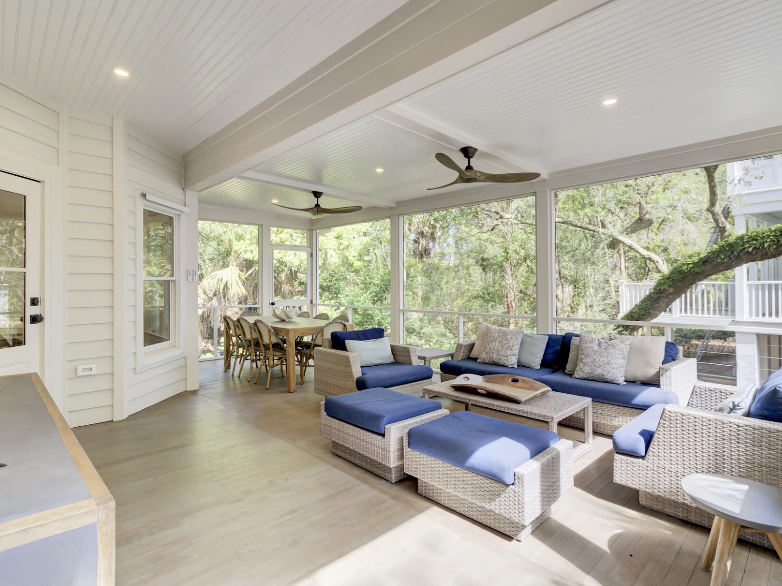 A bright, airy screened porch with white paneling, a sectional seating area in blue and gray, a ceiling fan, and a dining nook overlooking lush trees, embracing outdoor living.