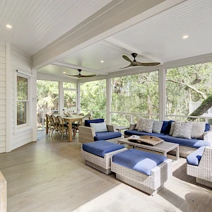 A bright, airy screened porch with white paneling, a sectional seating area in blue and gray, a ceiling fan, and a dining nook overlooking lush trees, embracing outdoor living.