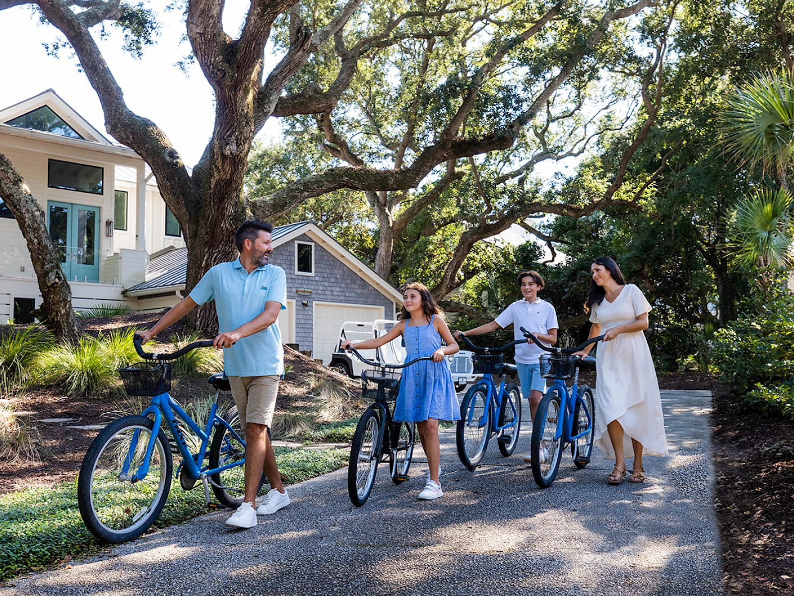 A family jogging bikes along a sunny path by a white house, with trees overhead and everyone smiling on ebikes.