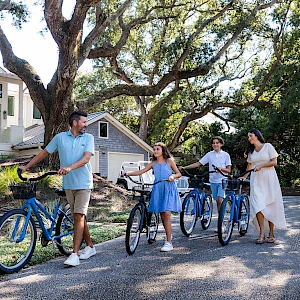 A family jogging bikes along a sunny path by a white house, with trees overhead and everyone smiling on ebikes.