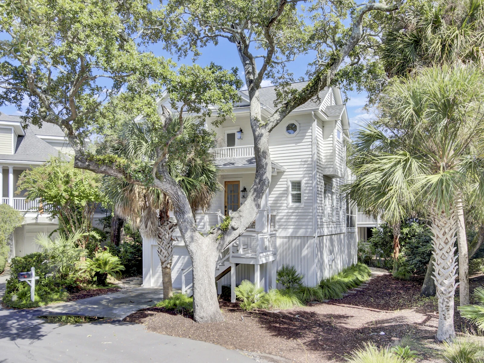 A two-story light-gray townhouse with a small front yard, trees, and shrubs; sunny day, palm trees on the sides, residential neighborhood.