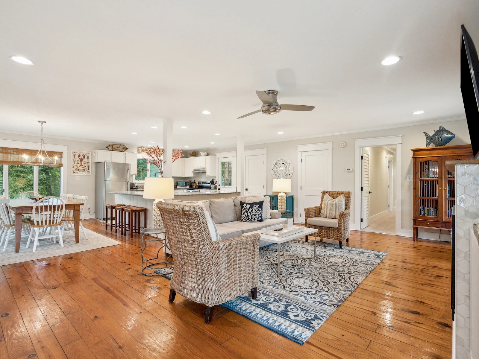 A bright, open-concept living and dining area with wooden floors, white walls, a blue rug, wicker chairs, a kitchen island, and a TV mounted on a wall.